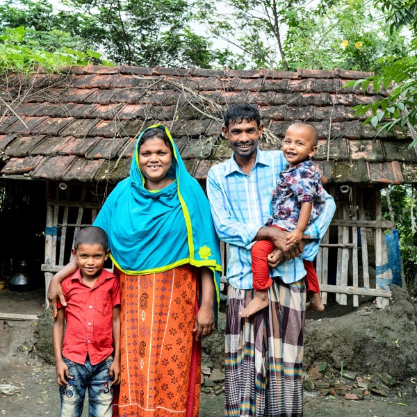 A mother and a father stand smiling with their two sons outside of their home in Bangladesh.