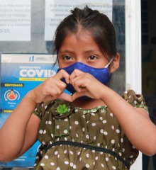 A young girl, wearing a mask, makes a heart with her hands.