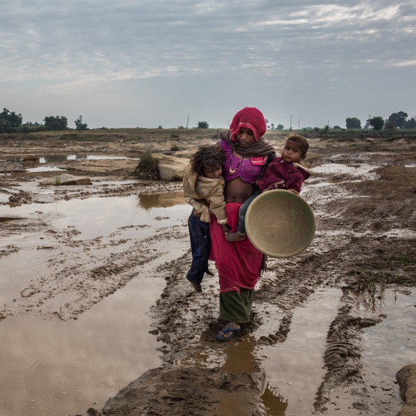 A mother carries her two children over a muddy road.
