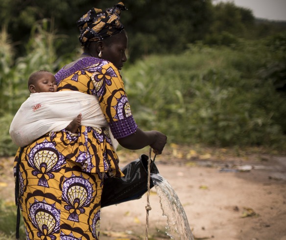 A mother, carrying a child on her back, pours clean water from a well into a bucket.